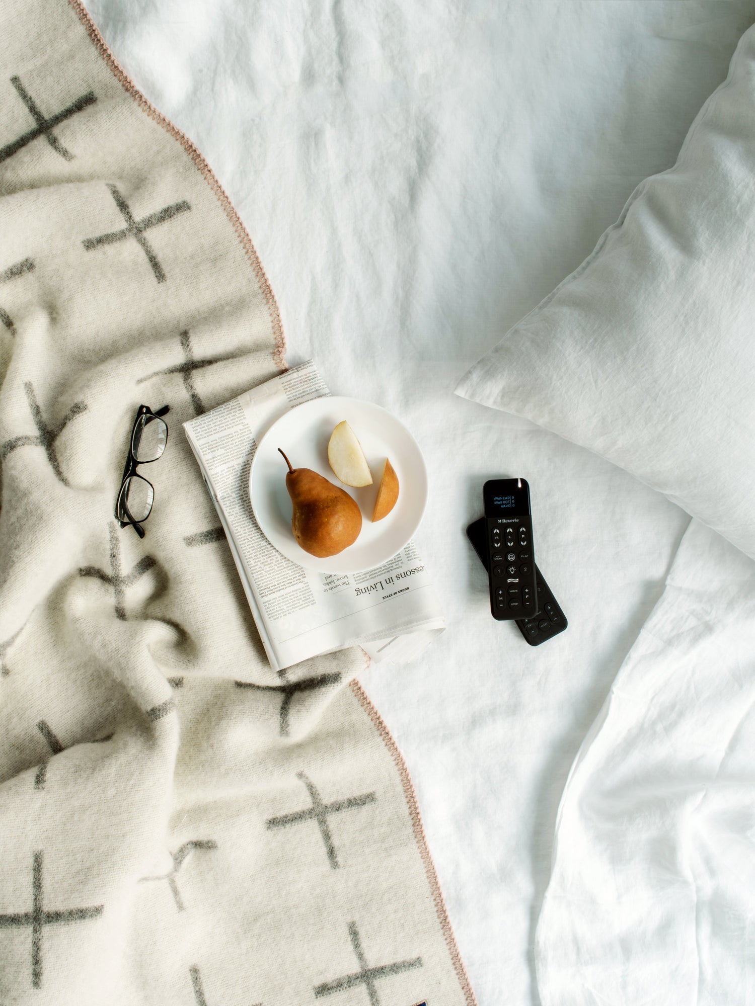 Two black adjustable bed remotes sitting next to a plate with a bosc pear on top of a magazine on top of a bed. 