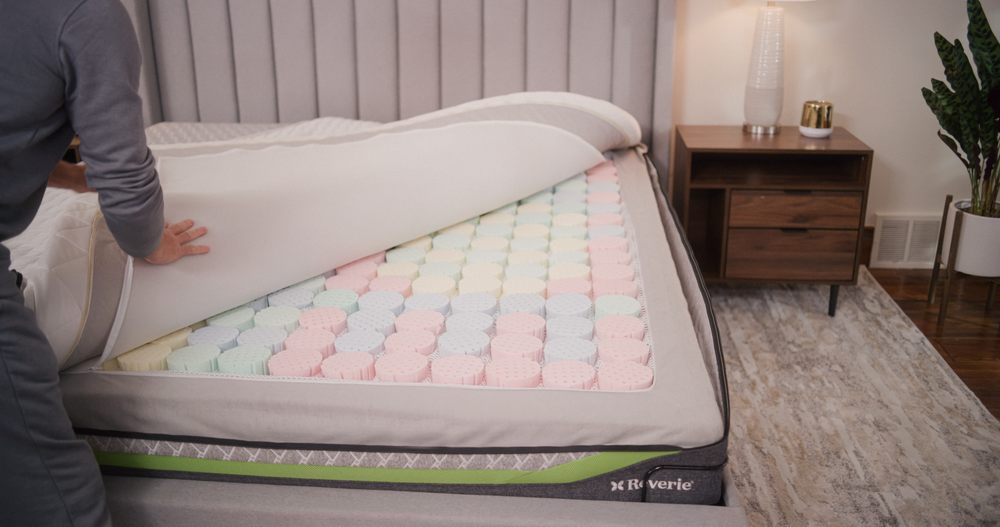 Person pulling back a mattress cover and topper on unzipped Reverie mattress to reveal DreamCell foam springs in a bedroom setting.