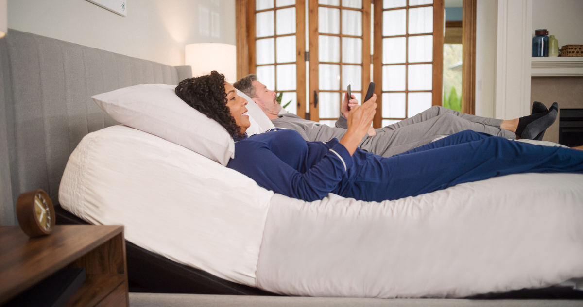 Two people lying on a white bean bag chair in a room with a lamp and window.