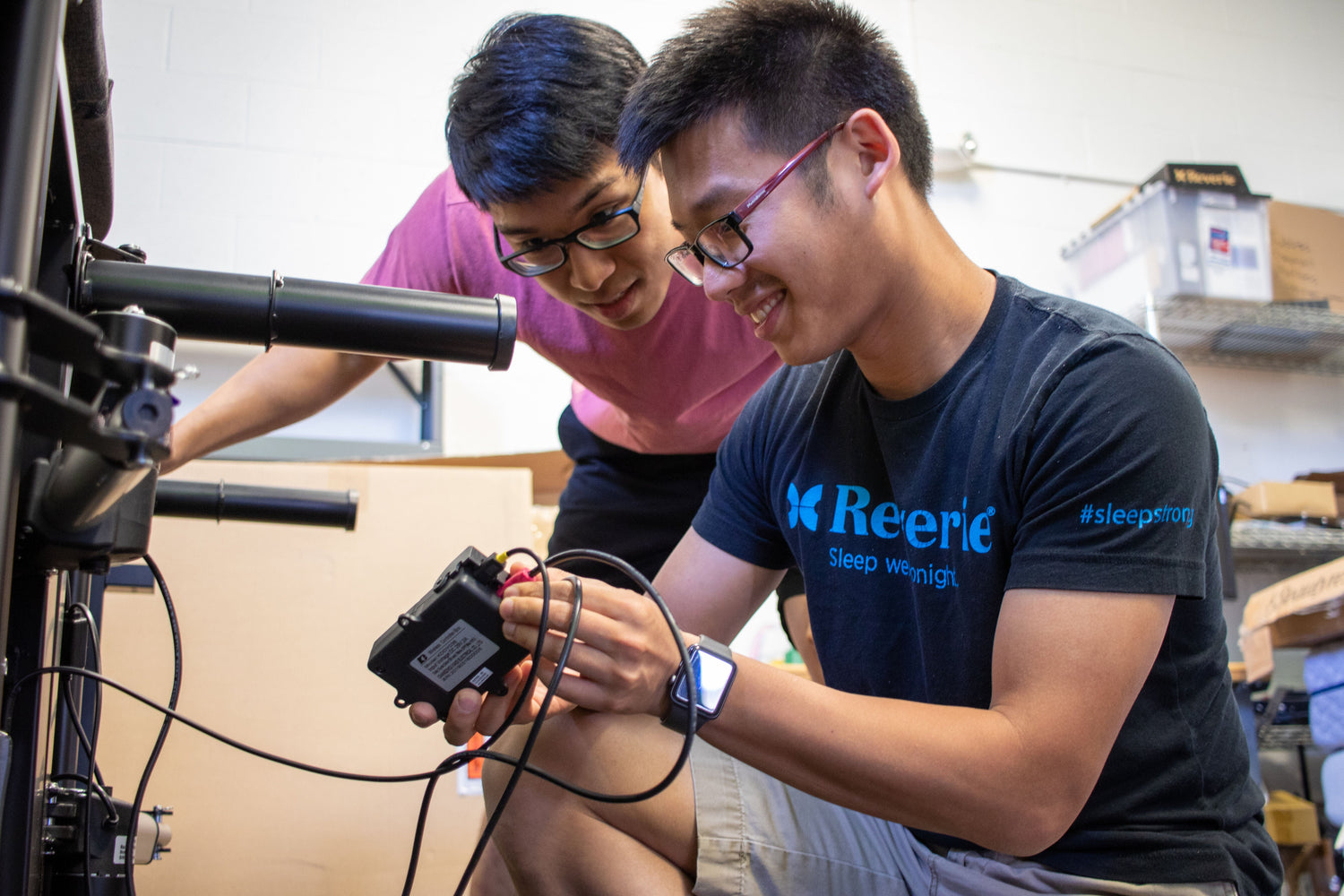 Two individuals working on a mechanical or electronic device in a workshop setting.