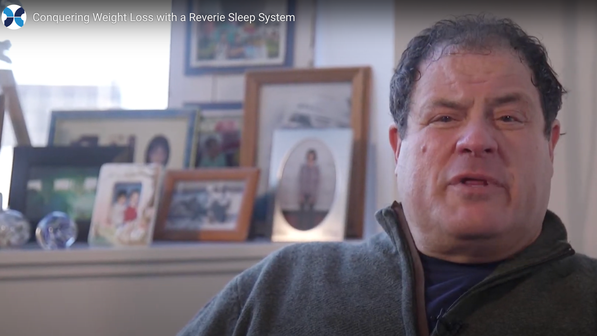 Man in a gray sweater speaking in front of a wall with framed photos