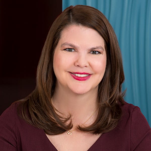 Woman with brown hair and red lipstick smiling in front of a blue curtain